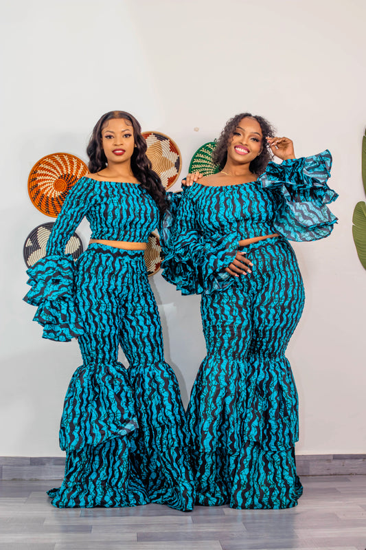 Two women in matching teal and black patterned outfits standing against a white wall.